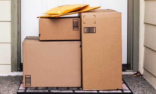 Three stacked cardboard boxes and two padded envelopes sit on a doormat at a front door. Represents home delivery and online shopping.